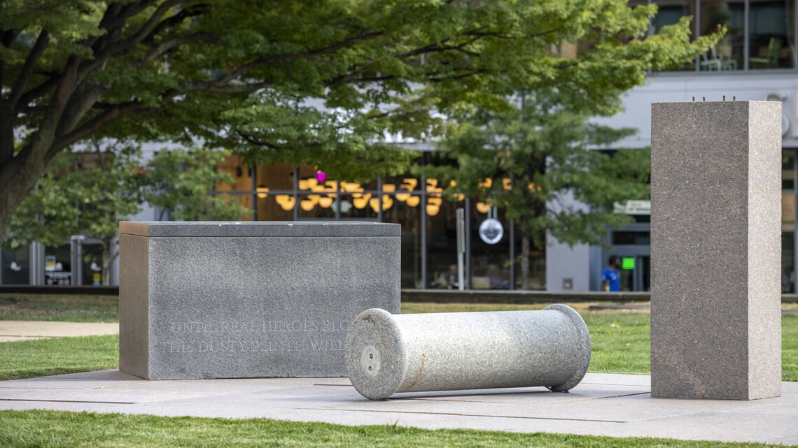 In the center, three sculptural objects in this order: a stone rectangular plint, a fallen column with rounded ends, and another column standing tall. All three are various shades of gray and rest on a base of concrete surrounded by green grass. In the background on the left, a tall tree and its branches fan out to the right.